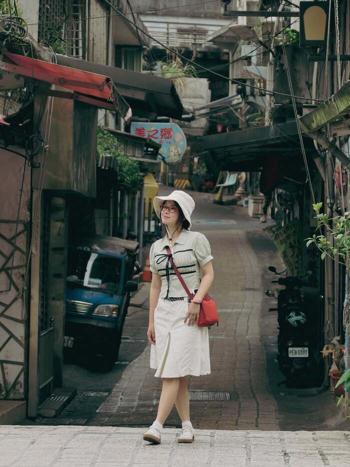 Hoàng Yến Chibi diện trang phục năng động với mũ bucket và túi đeo chéo đỏ, đang dạo bước giữa một con ngõ dốc cổ kính ở Taipei. Ảnh: Instagram nhân vật